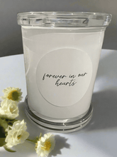 Load image into Gallery viewer, Forever In Our Hearts memorial candle in a clear glass jar, styled with small white flowers