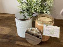 Load image into Gallery viewer, Champagne embossed soy candle jar with silver lid and product card, styled beside greenery on a wooden surface.
