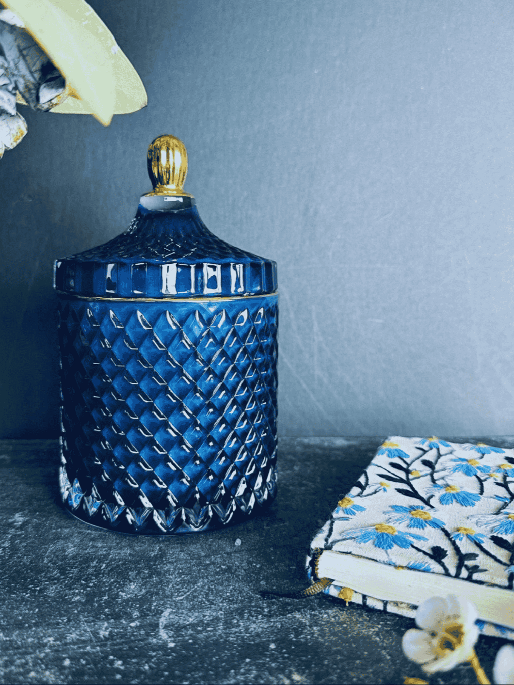 Deluxe royal blue geo soy candle with gold trim lid, styled on a dark surface beside a floral embroidered notebook.