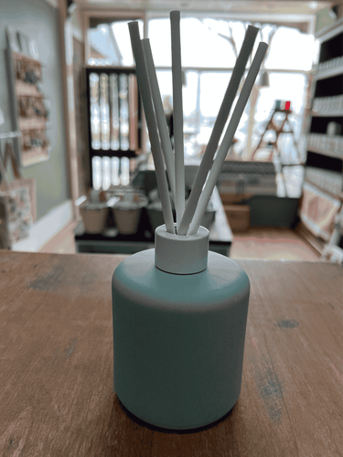 Sage Green matte reed diffuser with white reeds on display table.