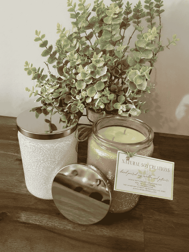 Luxury white embossed candle jar with soy wax and silver lid, displayed on a wooden surface beside greenery and product card.