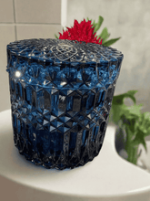 Load image into Gallery viewer, Midnight blue cut glass soy candle with intricate diamond pattern and matching lid, styled with greenery and red flower accent.
