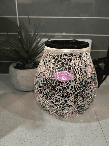 Mosaic electric wax melt warmer with pink and purple glass design, glowing light and fitted globe, displayed on a tiled surface beside a plant.