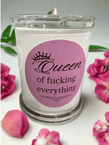 White glass candle with pink label that reads: Queen of fucking everything, styled with pink rose petals