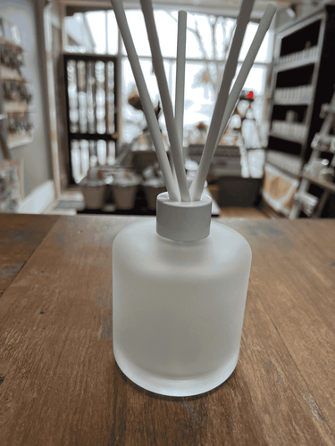Frosted glass reed diffuser bottle with white reeds on wooden table.