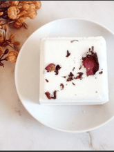 Load image into Gallery viewer, Natural Soy Creations rose geranium shower fizzy with pink petals on white plate beside dried flowers.
