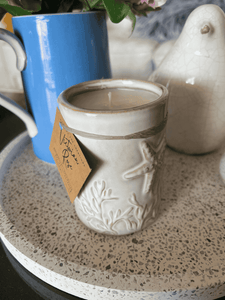 Natural Soy Creations Seaside soy candle in a white ceramic jar with seashell and starfish design on display with other items.
