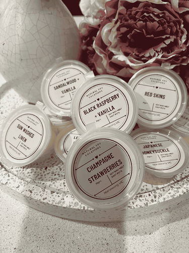 Assorted soy melt shot pots in fragrances including Black Raspberry Vanilla, Champagne Strawberries, Japanese Honeysuckle, Sandalwood Vanilla, Sun Washed Linen, Red Skins and Lemon, styled with flowers and décor.