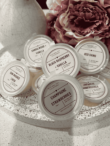 Assorted soy melt shot pots in fragrances including Black Raspberry Vanilla, Champagne Strawberries, Japanese Honeysuckle, Sandalwood Vanilla, Sun Washed Linen, Red Skins and Lemon, styled with flowers and decor.