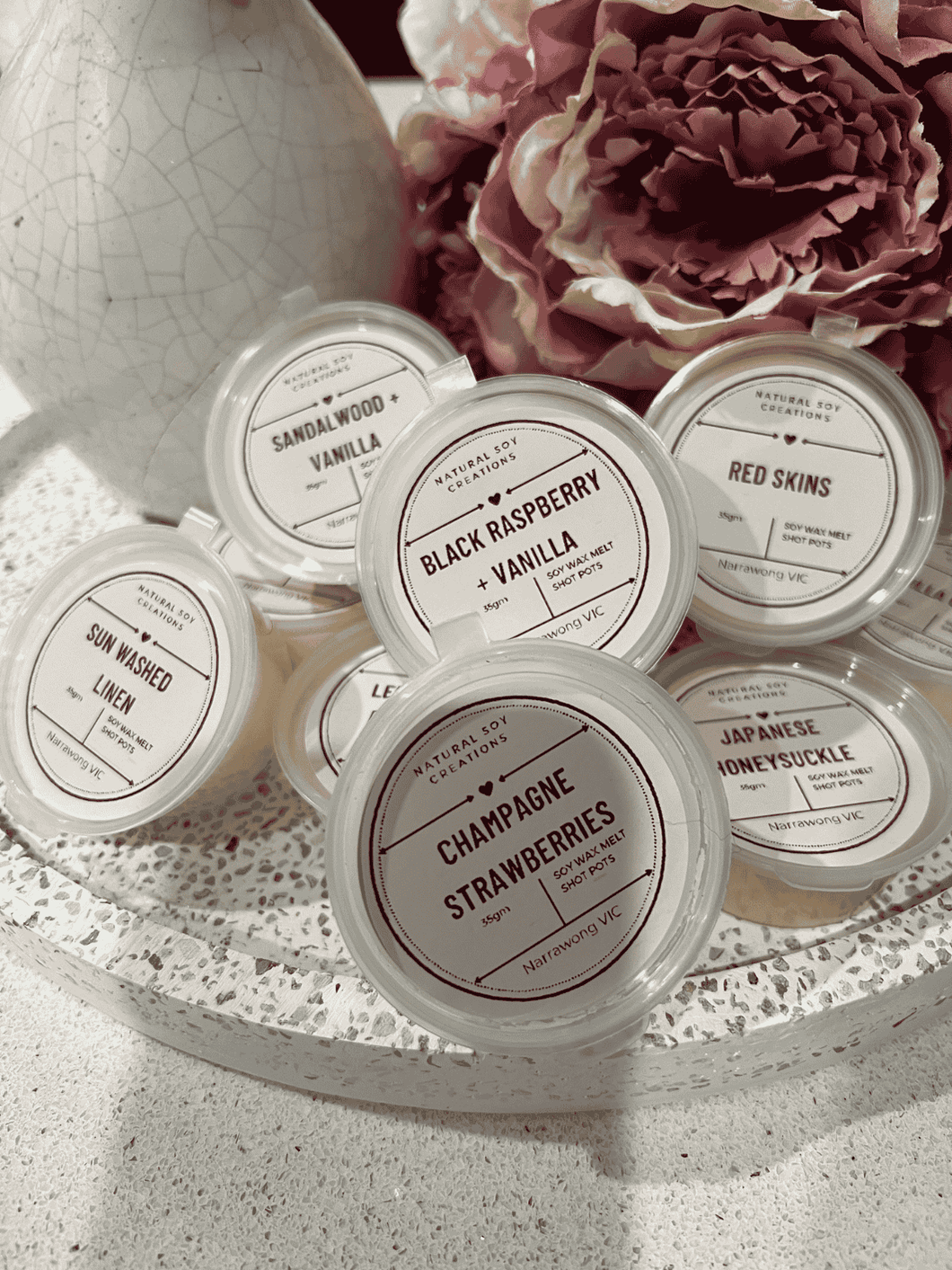 Assorted soy melt shot pots in fragrances including Black Raspberry Vanilla, Champagne Strawberries, Japanese Honeysuckle, Sandalwood Vanilla, Sun Washed Linen, Red Skins and Lemon, styled with flowers and decor.