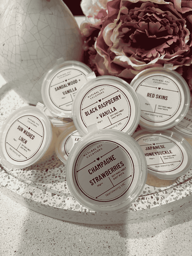 Assorted soy melt shot pots in fragrances including Black Raspberry Vanilla, Champagne Strawberries, Japanese Honeysuckle, Sandalwood Vanilla, Sun Washed Linen, and Red Skins, styled with flowers and decor.