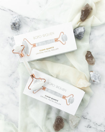 Clear quartz crystal facial roller by Bopo Women, displayed on marble with packaging and natural stones, promoting glowing and hydrated skin.