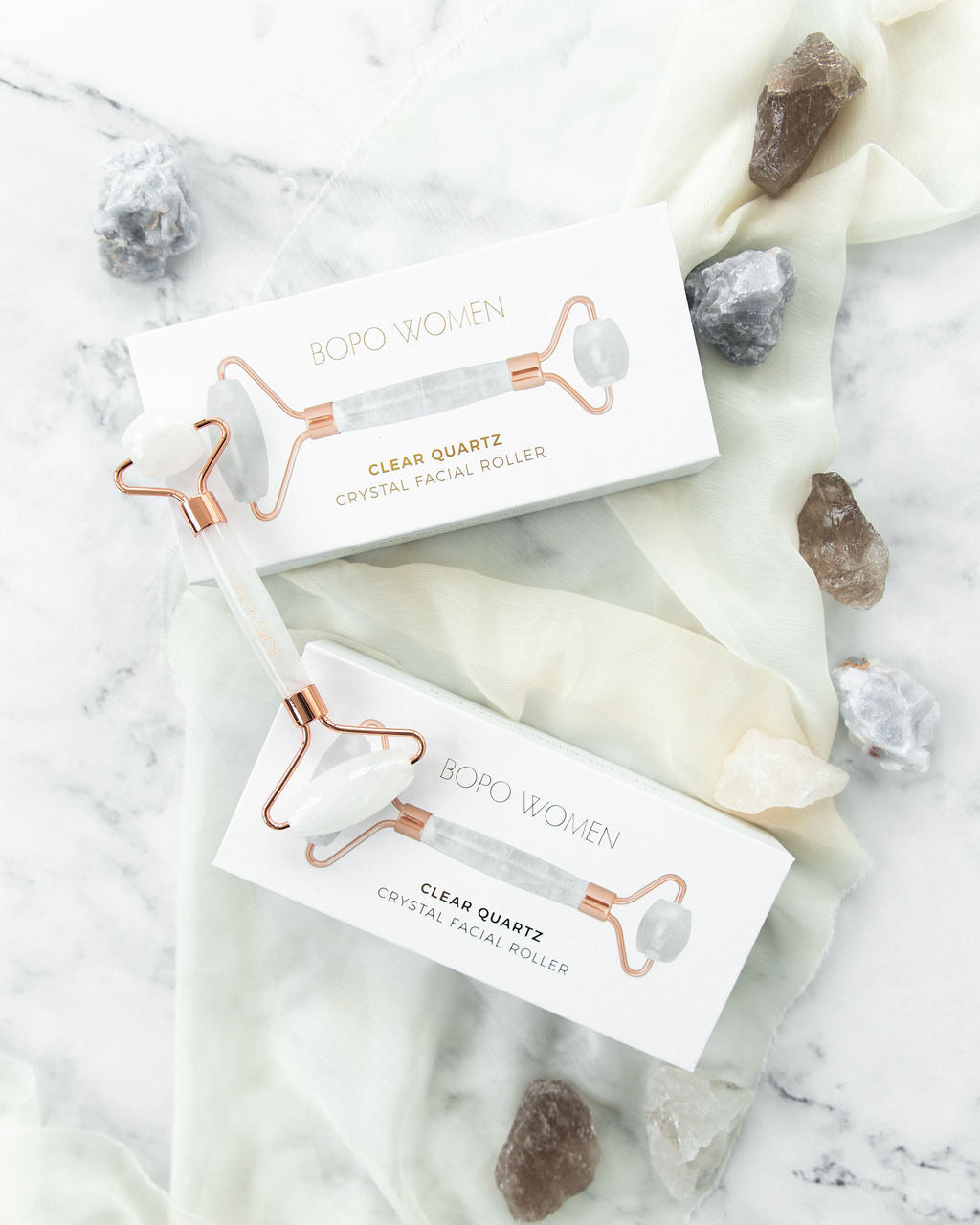 Clear quartz crystal facial roller by Bopo Women, displayed on marble with packaging and natural stones, promoting glowing and hydrated skin.