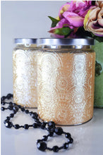 Load image into Gallery viewer, Champagne embossed candle jar with silver lid displayed beside flowers and black beaded necklace on a light background.
