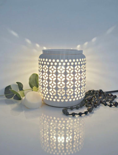 White ceramic electric wax melt warmer with decorative cut-out design, glowing softly beside black beaded decor and green leaves.